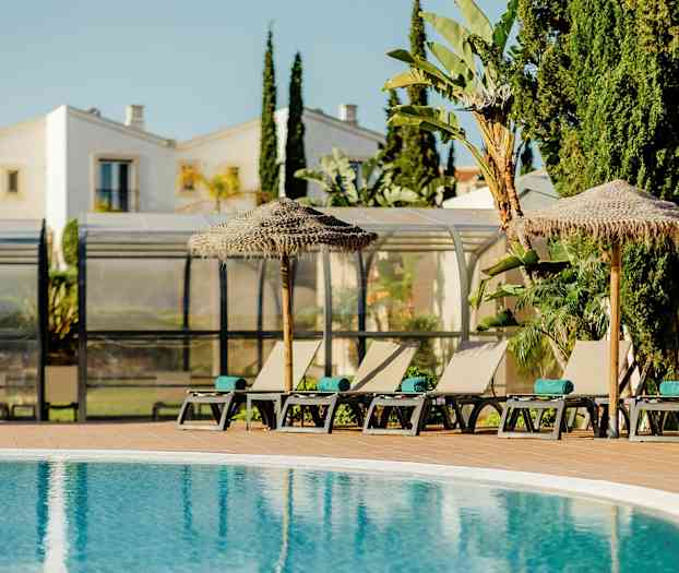 Aurinkotuoleja ja varjoja altaan vieressä, taustalla rakennuksia ja kasvillisuutta. (Tekoälyn luoma teksti)