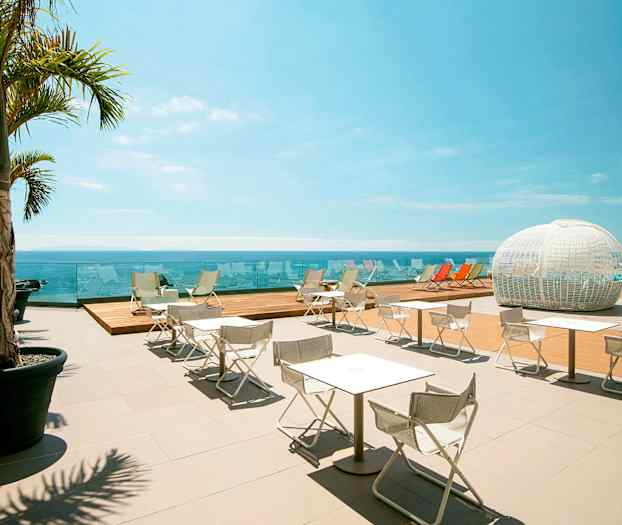 Ulkoterassi, jolla on valkoisia tuoleja ja pöytiä, varjostavia palmuja, näkymä merelle sekä valkoinen kupolimainen rakenne taustalla. (Tekoälyn luoma teksti)