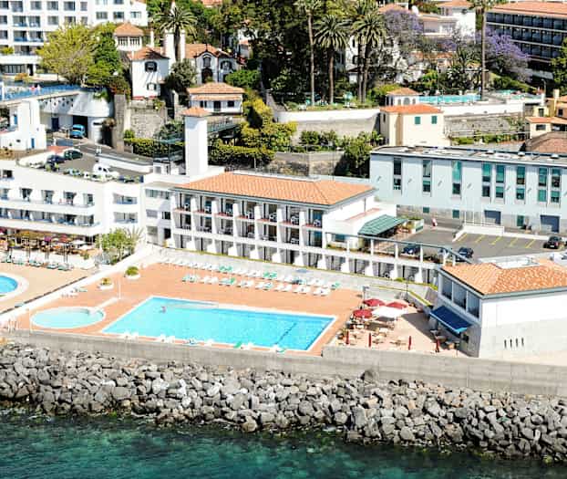 Ilmakuva hotellista, jossa on uima-altaita, aurinkotuoleja ja rakennuksia lähellä merta. (Tekoälyn luoma teksti)