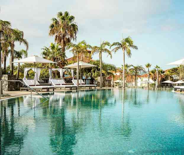 Ylellinen uima-allas aurinkotuoleineen ja päivänvarjoineen, ympärillä palmuja ja rakennuksia taustalla. (Tekoälyn luoma teksti)