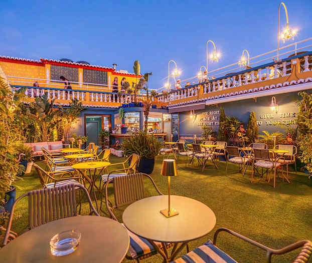 Ulkoterassi, jossa pöytiä ja tuoleja, koristeltu kasveilla ja valaistuksella. (Tekoälyn luoma teksti)