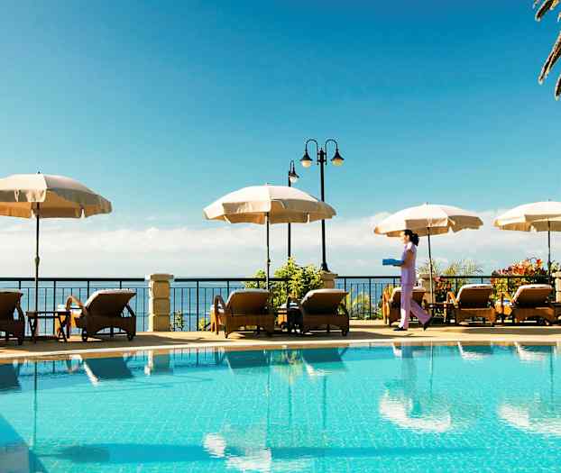 Ulkouima-allas, jonka vieressä aurinkotuoleja ja aurinkovarjoja sekä henkilö, joka kantaa tarjotinta. (Tekoälyn luoma teksti)