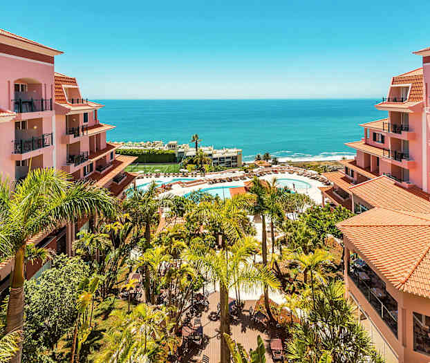 Näkymä hotellialueelle, jossa vaaleanpunaisia rakennuksia, palmuja ja uima-allas aurinkotuoleineen lähellä merta. (Tekoälyn luoma teksti)