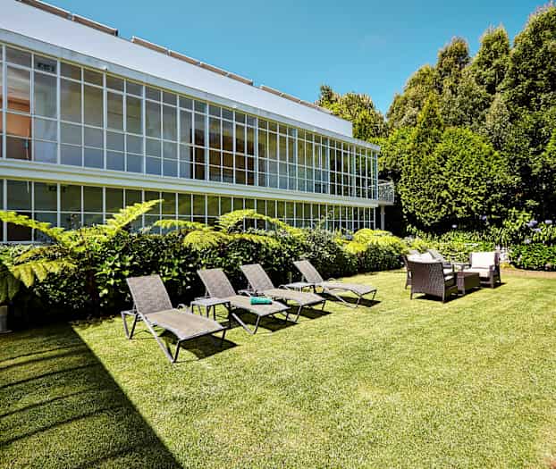 Kuva puutarhasta, jossa aurinkotuoleja, istuinryhmä ja vihreitä kasveja suuren ikkunarakennuksen edessä. (Tekoälyn luoma teksti)