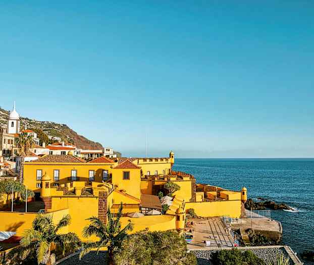 Keltainen rakennus meren rannalla, ympäröivänä vehreä alue ja mäkiä taustalla. (Tekoälyn luoma teksti)