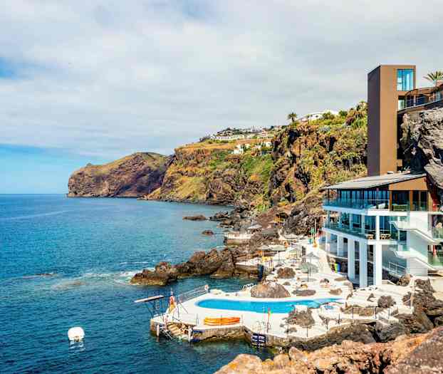 Rannikkomaisema, jossa on nykyaikainen rakennus ja uima-allasalue kallioiden laidalla, ympärillä meri ja vehreät rinteet. (Tekoälyn luoma teksti)