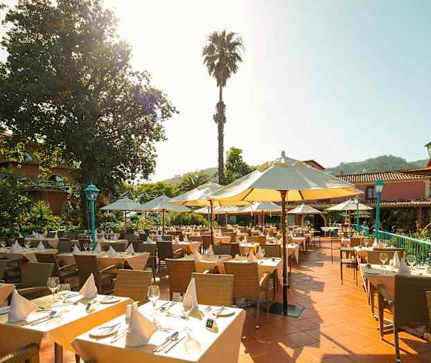 Ulkoterassi, jossa on katettuja pöytiä, aurinkovarjoja ja vehreä ympäristö. (Tekoälyn luoma teksti)