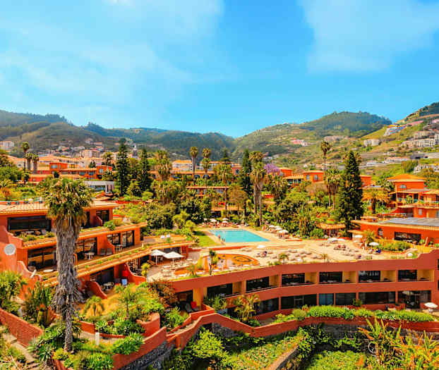 Rakennuskompleksi, jossa on terrakotanvärisiä rakennuksia, uima-allas ja vehreää kasvillisuutta, joka sijaitsee laaksossa mäkien ympäröimänä. (Tekoälyn luoma teksti)