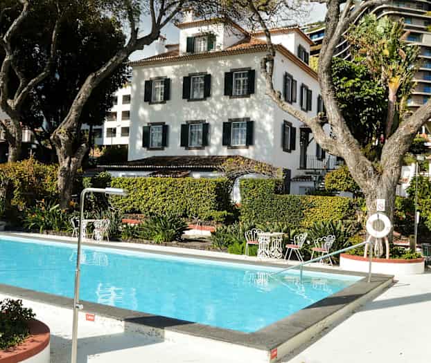 Kuva ulkouima-altaasta, jossa vettä, ympärillä puita ja pensaita, taustalla valkoisia rakennuksia. (Tekoälyn luoma teksti)