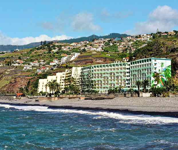 Hotellirakennuksia rannalla, jossa on meren aaltoja ja taustalla vihreitä kukkuloita sekä asuinrakennuksia. (Tekoälyn luoma teksti)