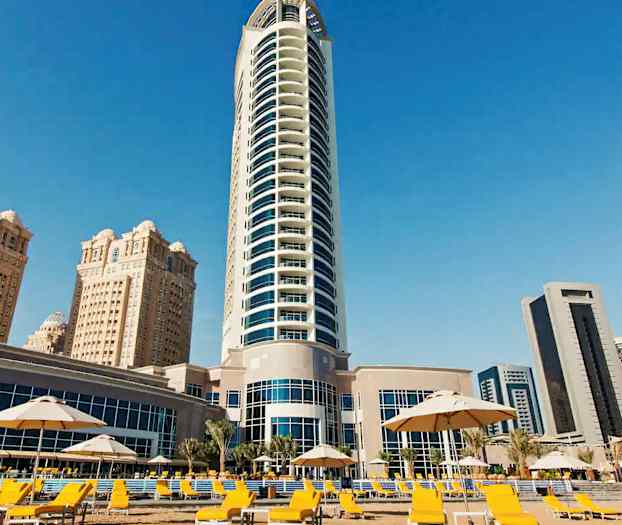 Kuvassa näkyy moderni korkea rakennus, jota ympäröivät useat muut rakennukset ja alue, jossa on keltaisia aurinkotuoleja ja varjoja. (Tekoälyn luoma teksti)