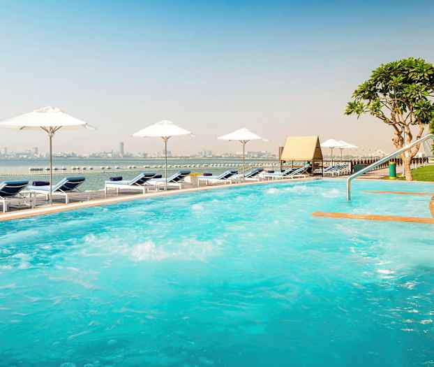 Ulkouima-allas aurinkotuoleilla ja aurinkovarjoilla meren rannalla, kaupungin siluetti näkyy taustalla. (Tekoälyn luoma teksti)