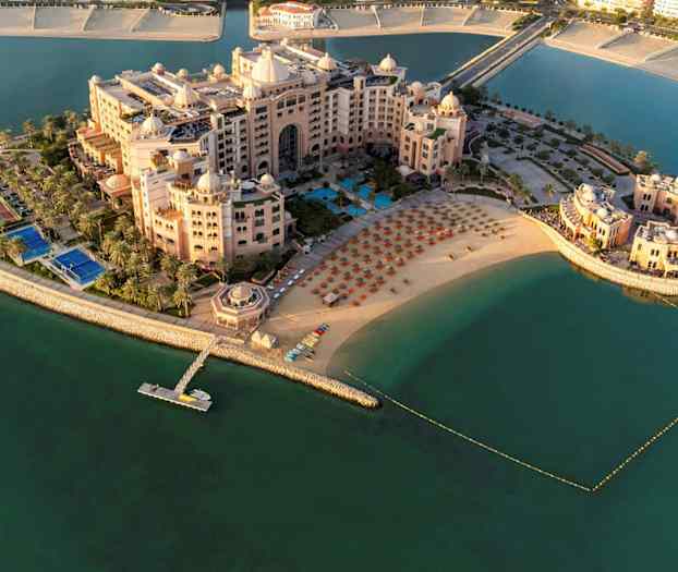 Ilmakuva ylellisestä hotellikompleksista rannalla, jossa on aurinkotuoleja ja varjoja, veden ympäröimänä. (Tekoälyn luoma teksti)