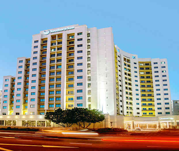 Moderni hotelli, jossa on korkea rakennus ja paljon ikkunoita, sijaitsee vilkkaasti liikennöidyn kadun vieressä illalla. (Tekoälyn luoma teksti)