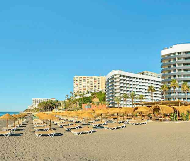 Ranta, jolla on aurinkotuoleja ja aurinkovarjoja, ympärillä useita hotellirakennuksia. (Tekoälyn luoma teksti)