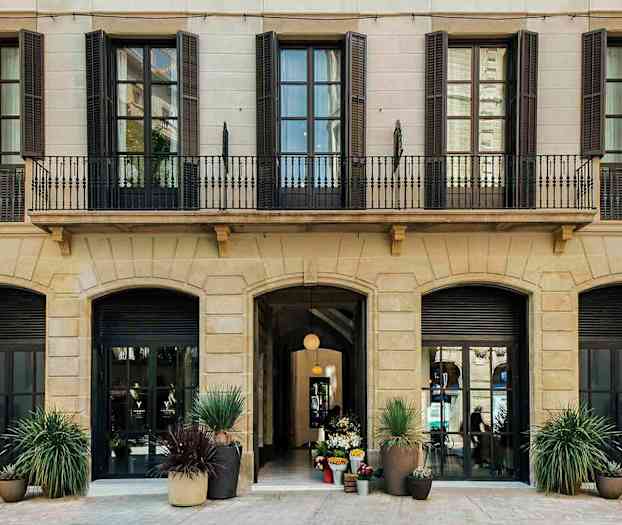 Kuva rakennuksen julkisivusta, jossa on useita korkeita ikkunoita ja sisäänkäynti koristeltuna kukkaruukuilla. (Tekoälyn luoma teksti)