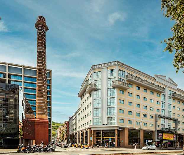 Kuva kaupunkimaisemasta, jossa on moderneja rakennuksia, kaksi korkeaa savupiippua sekä ihmisiä ja ajoneuvoja etualalla. (Tekoälyn luoma teksti)