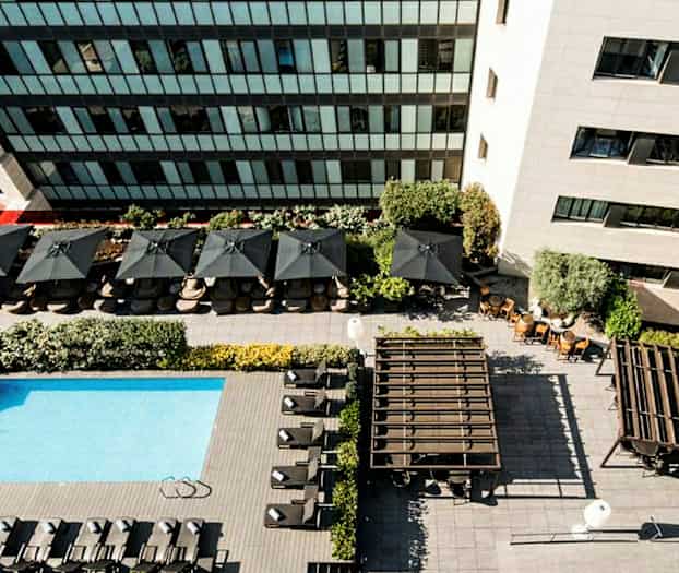 Ilmakuva uima-altaasta, jossa on aurinkotuoleja, aurinkovarjoja ja ympäröiviä rakennuksia. (Tekoälyn luoma teksti)