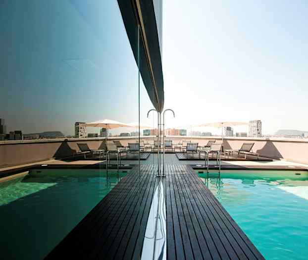 Kattouima-allas puualustoilla ja aurinkotuoleilla vieressä. Kaupunki näkyy taustalla. (Tekoälyn luoma teksti)