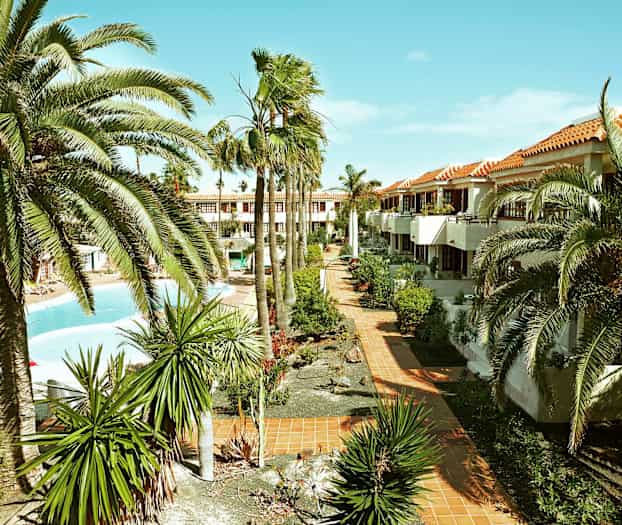 Kuva lomakohteesta, jossa on palmuja, puutarhoja ja uima-allas. Rakennuksia, joissa on terrakotanväriset katot, näkyy taustalla. (Tekoälyn luoma teksti)