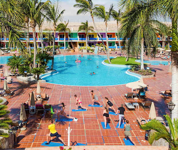 Suuren hotellin vieressä, jossa on värikkäitä rakennuksia, ihmiset harjoittavat ulkoilmajoogaa matoilla altaan äärellä. (Tekoälyn luoma teksti)