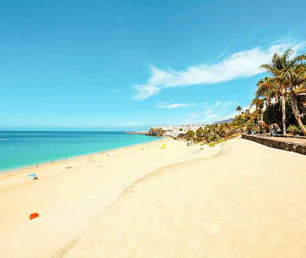 Pitkä hiekkaranta, jossa kirkasta sinistä vettä ja ihmisiä aurinkoa ottamassa ja uimassa. Rannan vieressä kulkee kävelytie, jolla on palmuja ja kasvillisuutta. (Tekoälyn luoma teksti)