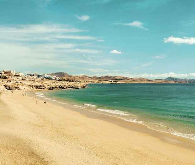 Leveä hiekkaranta, joka ulottuu pitkin rantaviivaa turkoosinvärisen veden ja taustalla näkyvien rakennusten kanssa. (Tekoälyn luoma teksti)