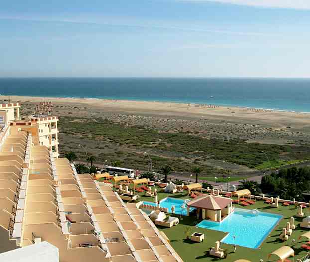 Kuva hotellialueelta, jossa etualalla uima-allas, aurinkotuoleja ja varjoja, ja taustalla pitkä hiekkaranta meren äärellä. (Tekoälyn luoma teksti)