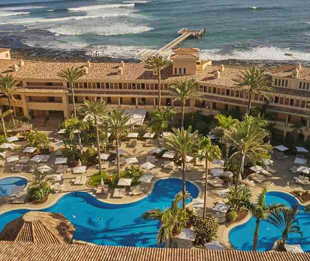 Hotellialue uima-altailla, aurinkotuoleilla ja aurinkovarjoilla, joiden ympärillä palmupuita. Taustalla näkyy meri aaltoineen ja laituri. (Tekoälyn luoma teksti)