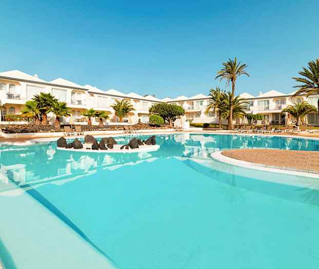 Kuva hotellin uima-altaasta, jossa on kirkasta sinistä vettä. Altaan ympärillä on palmuja ja valkoisia rakennuksia taustalla. (Tekoälyn luoma teksti)