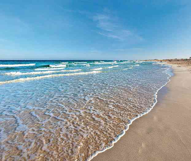 Hiekkaranta, jolla pehmeitä aaltoja huuhtoutuu, kirkkaan sinisen taivaan alla. (Tekoälyn luoma teksti)