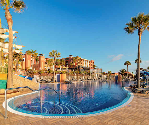 Ulkouima-allas aurinkoterassin kanssa, ympäröivät palmupuut ja taustalla rakennus. Etualalla näkyy koristeena laiva. (Tekoälyn luoma teksti)