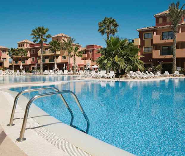 Lomakohde, jossa ulkouima-allas, ympärillä aurinkotuoleja ja palmuja sekä rakennuksia taustalla. (Tekoälyn luoma teksti)