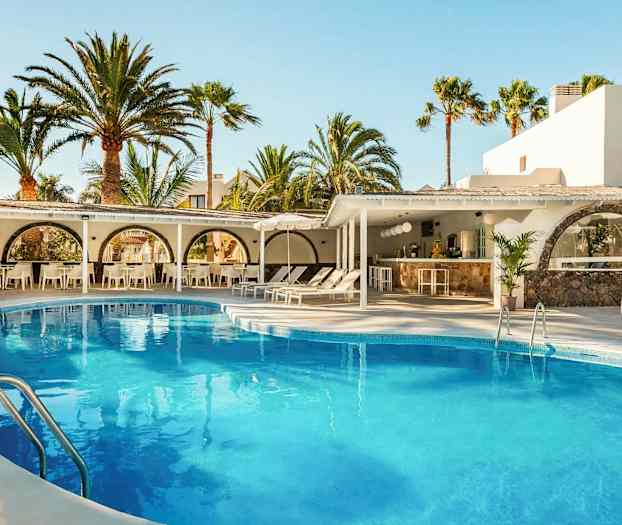 Ulkouima-allas, jota ympäröivät aurinkotuolit ja aurinkovarjot, taustalla valkoisia rakennuksia ja korkeita palmuja. (Tekoälyn luoma teksti)