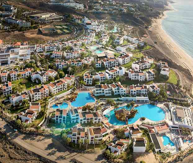 Ilmakuva lomakohteesta, jossa on valkoisia rakennuksia ja punaisia kattoja sekä suuria uima-altaita ja puutarhoja lähellä rantaa. (Tekoälyn luoma teksti)