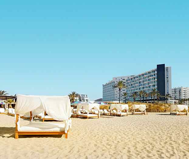 Ranta sänkyillä, joissa on valkoiset verhot; taustalla moderni rakennus. (Tekoälyn luoma teksti)