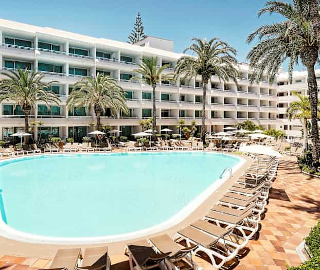 Hotellin uima-allas aurinkotuoleineen ja varjoineen, jota ympäröivät vehreät palmut, taustalla monikerroksinen rakennus parvekkeineen. (Tekoälyn luoma teksti)