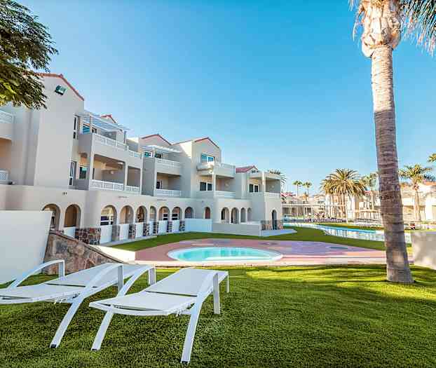 Ulkotila, jossa aurinkotuoleja, nurmikko, uima-allas ja lähellä oleva vaaleansävyinen rakennus. (Tekoälyn luoma teksti)