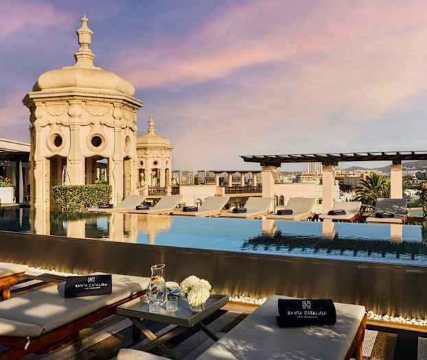 Kattoterassi, jossa on uima-allas ja aurinkotuoleja, taustalla kaupunkinäkymä ja koristeellinen arkkitehtuuri. (Tekoälyn luoma teksti)