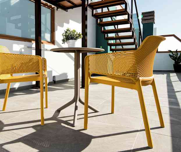 Kaksi keltaista tuolia ja pöytä aurinkoisella terassilla, jossa portaat ja kasvi. (Tekoälyn luoma teksti)