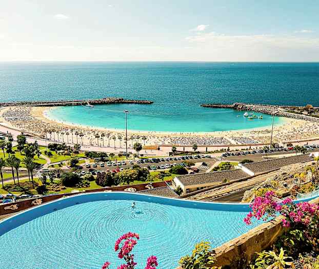 Ranta, jossa on kultaista hiekkaa ja kirkkaan sininen meri, ympäröity suojaisalla aallonmurtajalla ja vehreydellä. Etualalla uima-allas, jota koristavat kukat. (Tekoälyn luoma teksti)