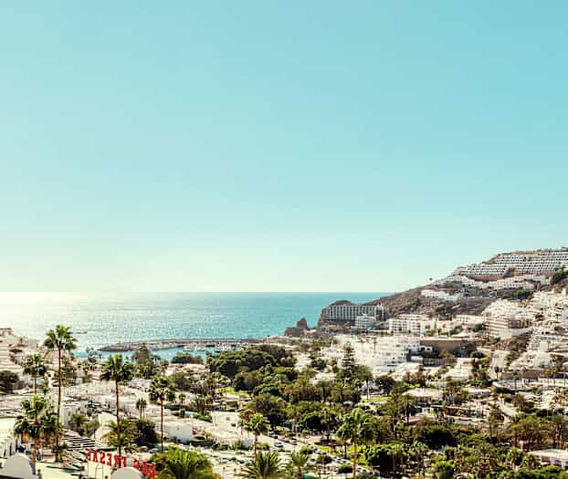 Näkymä rannikkokaupungista, jossa on valkoisia rakennuksia ja palmuja kimaltavan meren edessä. (Tekoälyn luoma teksti)