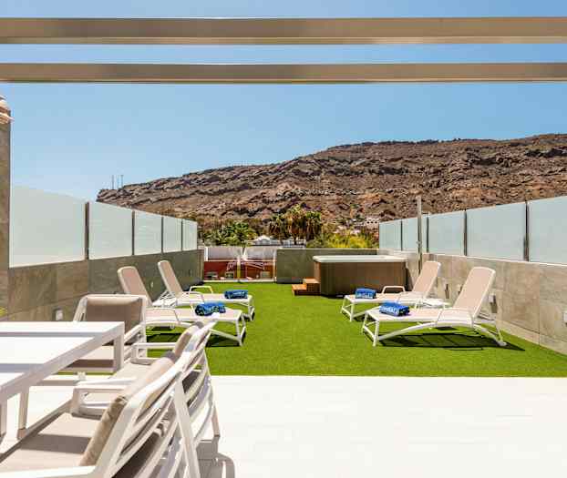 Ulkoilmaterassi, jossa on aurinkotuoleja tekonurmella ja poreallas, jota ympäröivät lasikaiteet ja vuoret taustalla. (Tekoälyn luoma teksti)