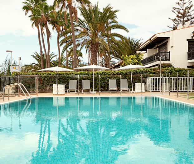 Kuva ulkouima-altaasta, jota ympäröivät aurinkotuolit ja aurinkovarjot, taustalla palmuja ja rakennus. (Tekoälyn luoma teksti)