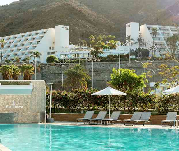 Uima-allas aurinkotuoleineen ja aurinkovarjoineen, taustalla valkoinen lomakeskus ja vuoria. (Tekoälyn luoma teksti)