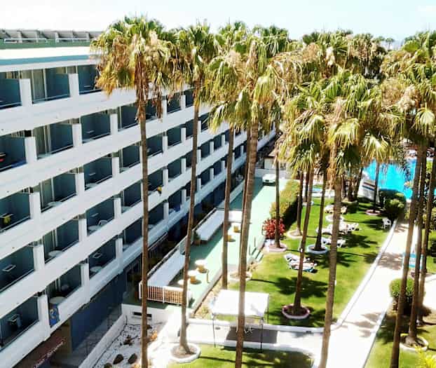 Hotelli, jossa on valkoiset julkisivut, ympäröitynä nurmikkoa palmuilla ja uima-altaalla. (Tekoälyn luoma teksti)