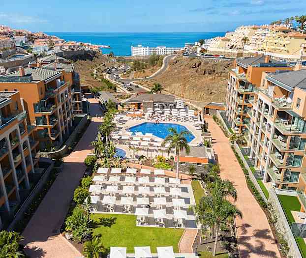 Hotellialue, jossa uima-allas, aurinkotuoleja ja ympäröiviä rakennuksia, taustalla meri. (Tekoälyn luoma teksti)