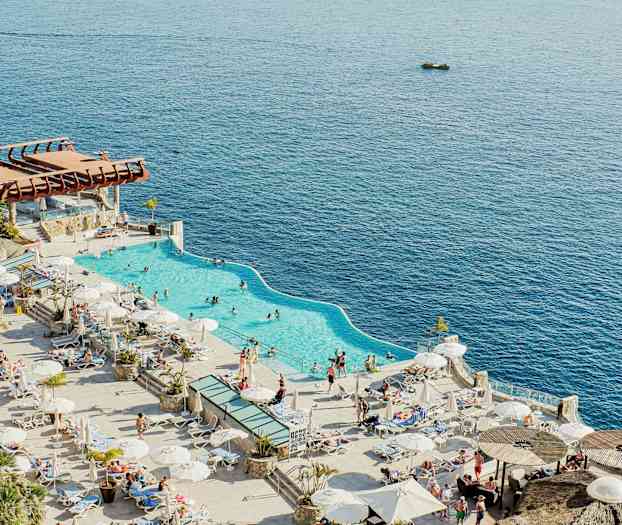 Kuva rantaläheisestä uima-altaasta aurinkotuolien ja varjojen ympäröimänä, taustalla meri. (Tekoälyn luoma teksti)