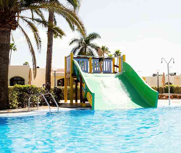 Vesiliukumäki uima-altaan vieressä, ympärillä palmuja ja rakennuksia. (Tekoälyn luoma teksti)