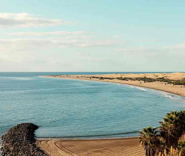 Näkymä rannalle, jossa hiekkadyynejä, meri ja kapea kivinen aallonmurtaja. (Tekoälyn luoma teksti)
